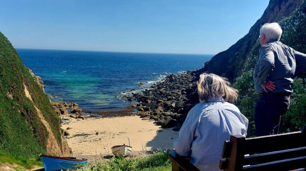 An older couple look out to sea above a Cornish Cove on a sunny day with blue sky, deep blue sea and fishing boats pulled up from the sandy beach