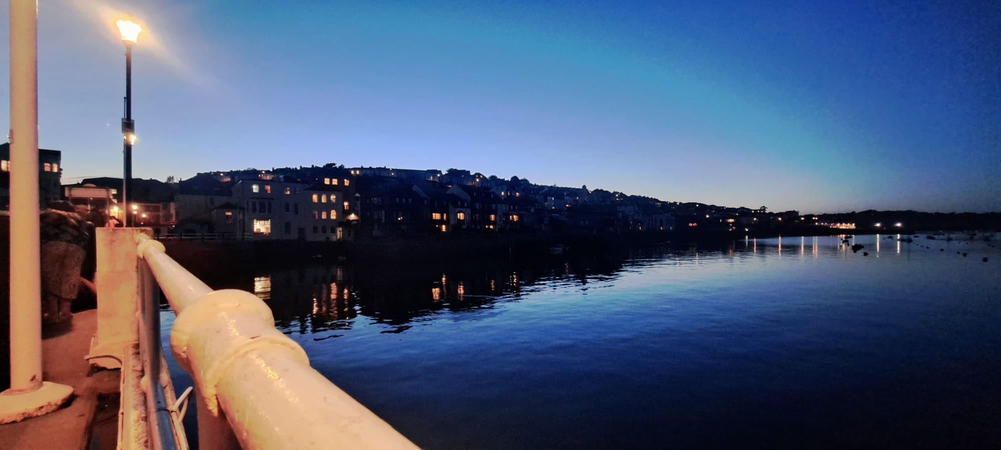 Lights of a harbour town at dusk as seen from a quay with railings and lights..