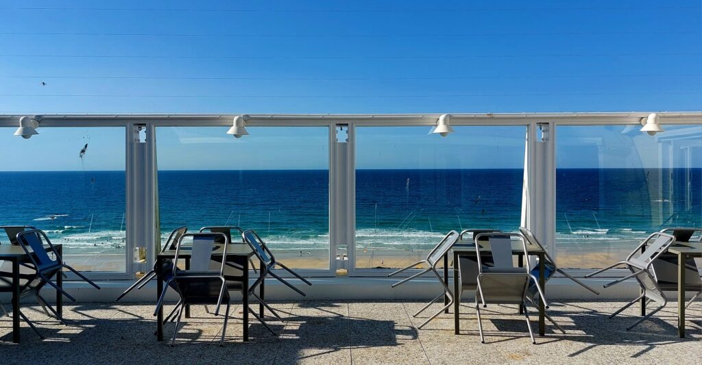 a view over the deep blue ocean from a cafe on top of the Tate Gallery in St Ives on a sunny day with sea blue sky. Chairs are leaned against tables.