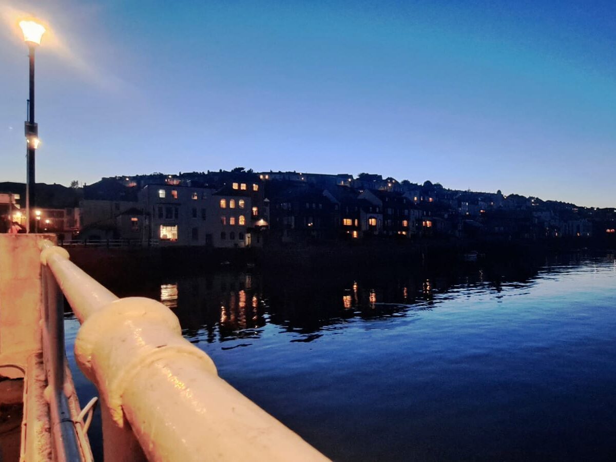 Lights of a harbour town at dusk as seen from a quay with railings and lights..